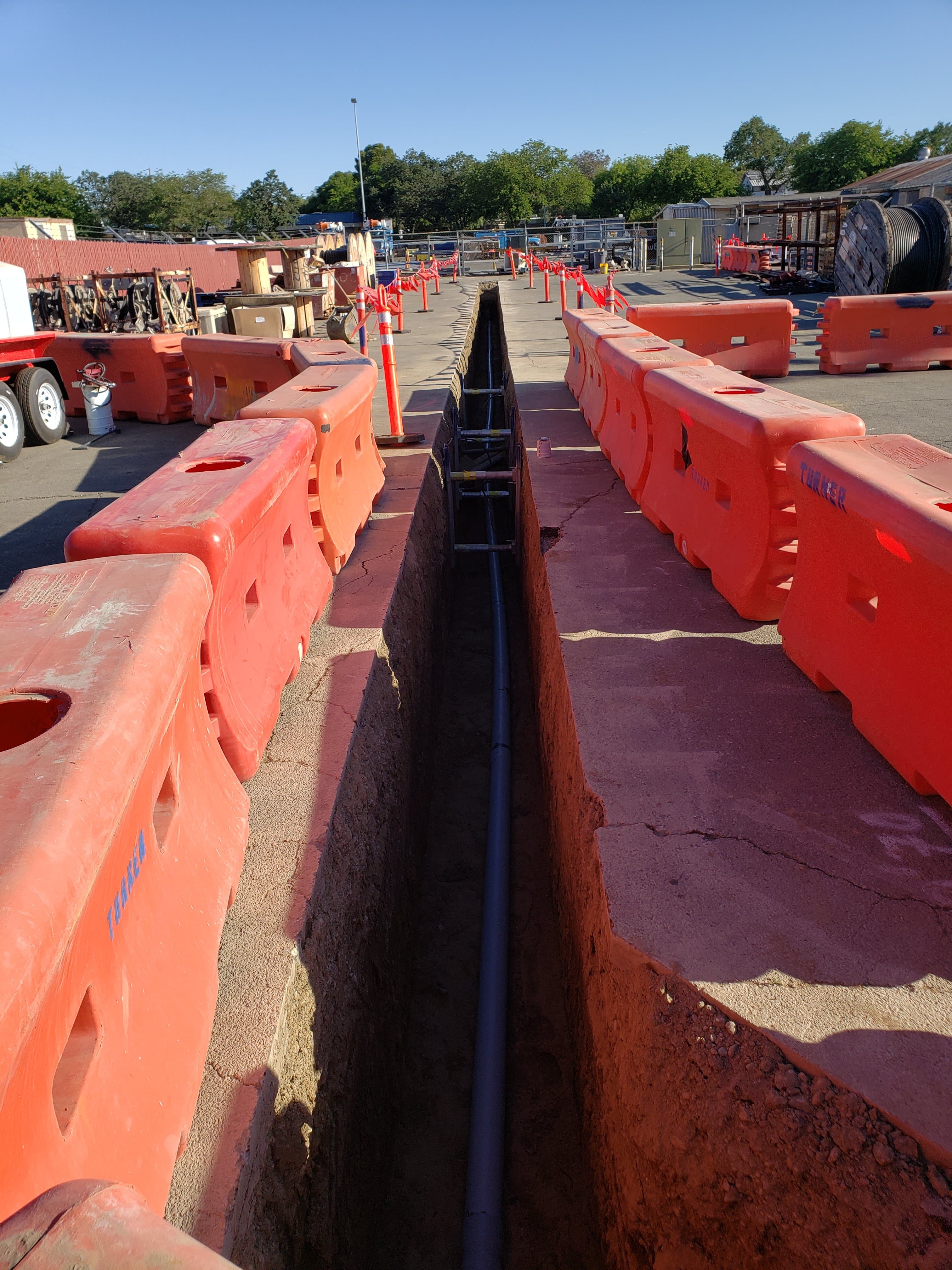 Underground conduit trench across a commercial equipment yard protected by orange water barriers